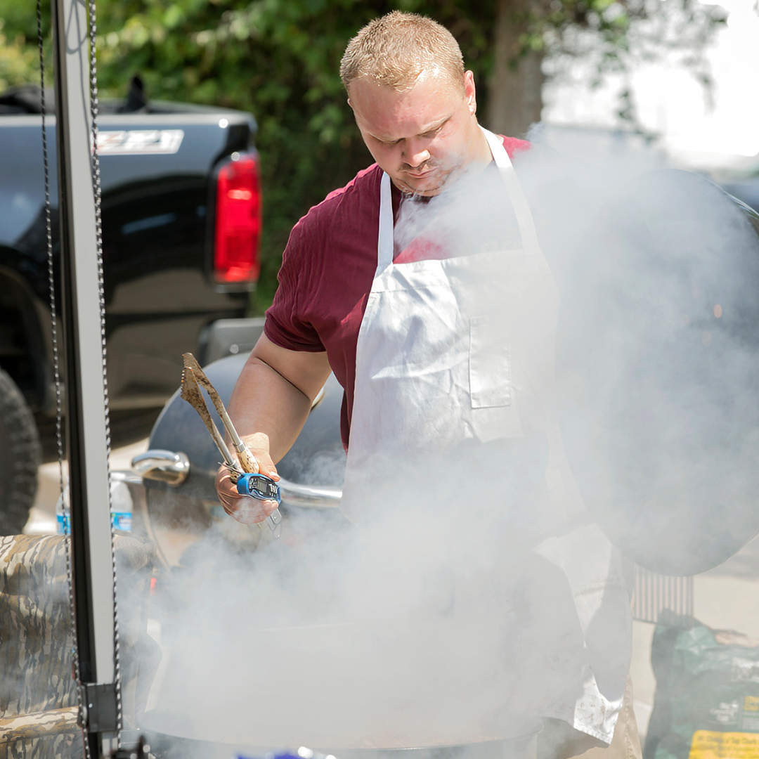 Man standing up and looking down as he is cooking over a smokey barbeque grill.