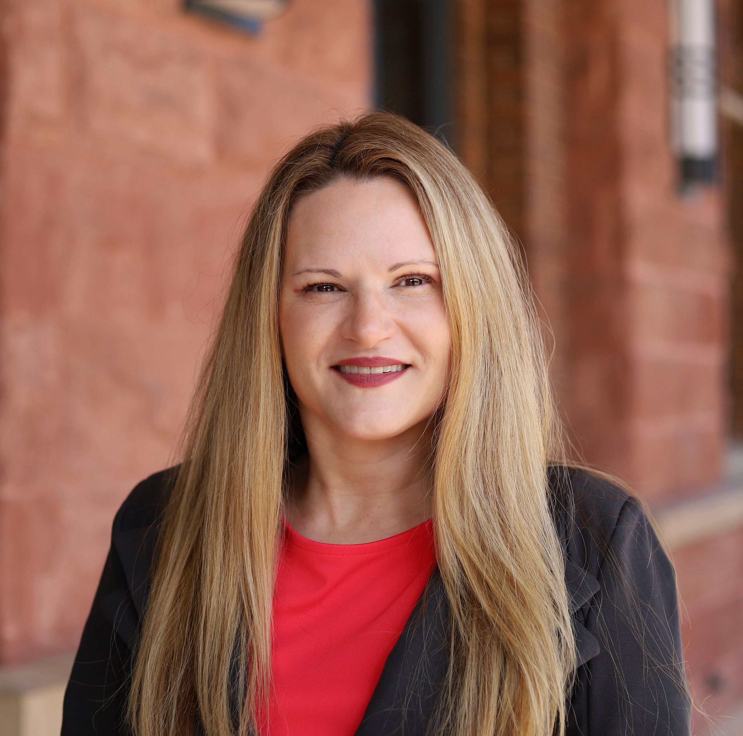 female with blonde hair wearing a red shirt and dark sport coat in front of red brick building 