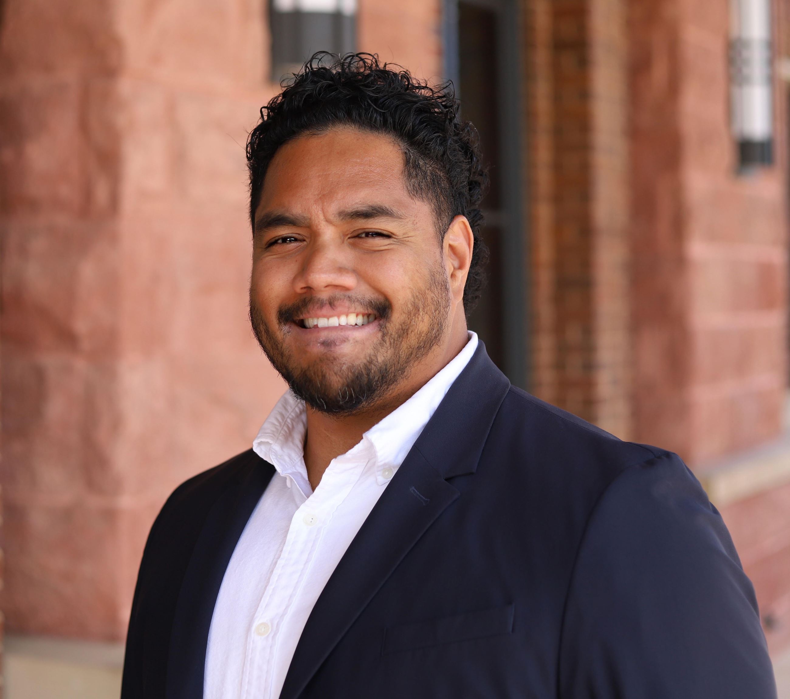man with short curly black hair wearing a white button up with a dark sport coat smiling 